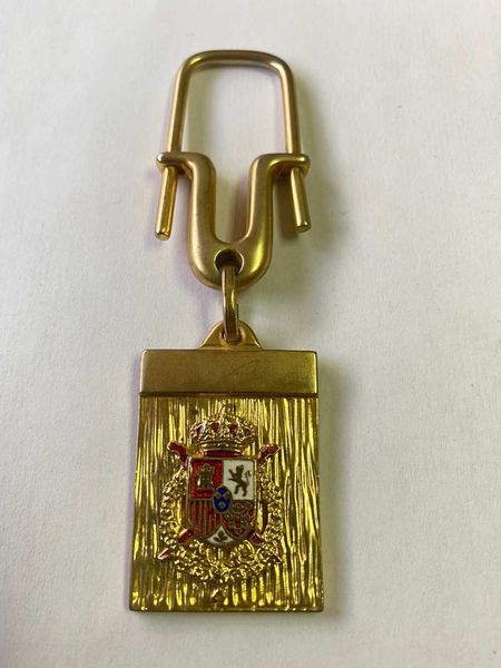 Llavero dorado con un colgante rectangular que luce el escudo de armas español.