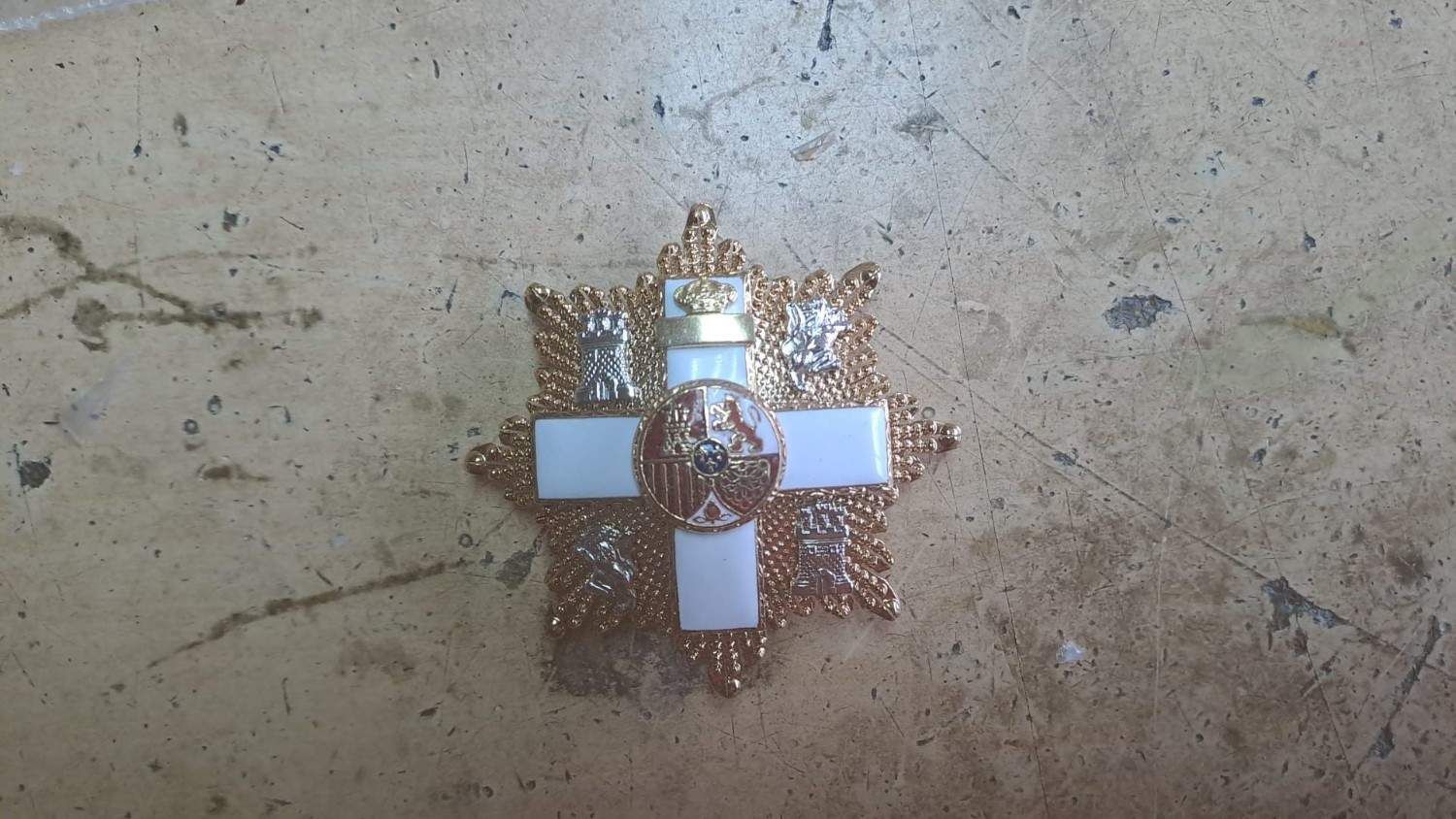 Medalla en forma de cruz blanca y dorada con una corona, sobre una superficie de piedra color canela.