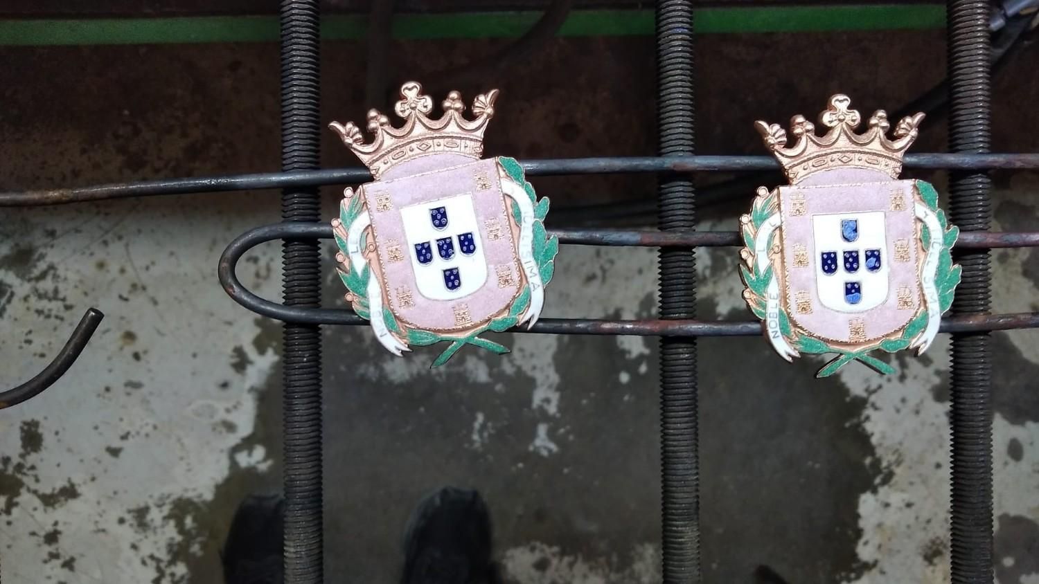 Dos escudos coronados de estilo portugués montados sobre una valla de metal desgastada.