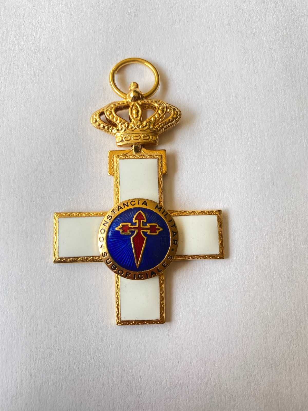 Colgante de cruz de oro con una corona y un emblema azul y rojo sobre un fondo blanco.