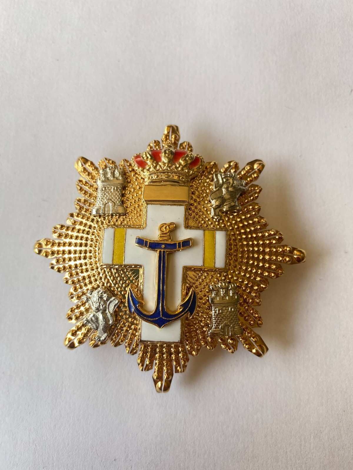 Ancla dorada y emblema de laurel con una cruz azul sobre fondo blanco