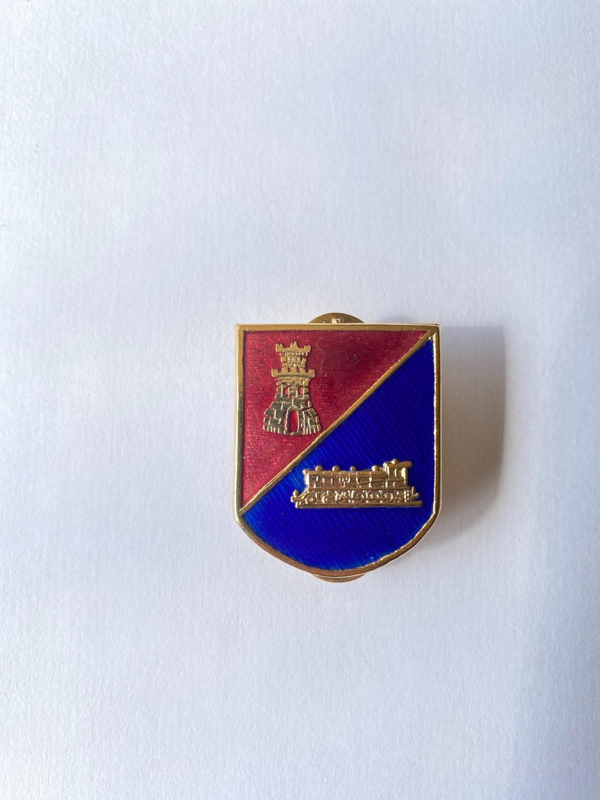 Pequeño pin esmaltado en forma de escudo, de color rojo y azul, con detalles dorados sobre fondo blanco.