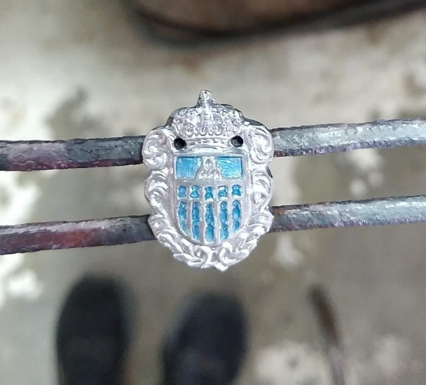 Primer plano de un pequeño emblema plateado con forma de escudo y detalles azules, sujeto con pinzas metálicas.