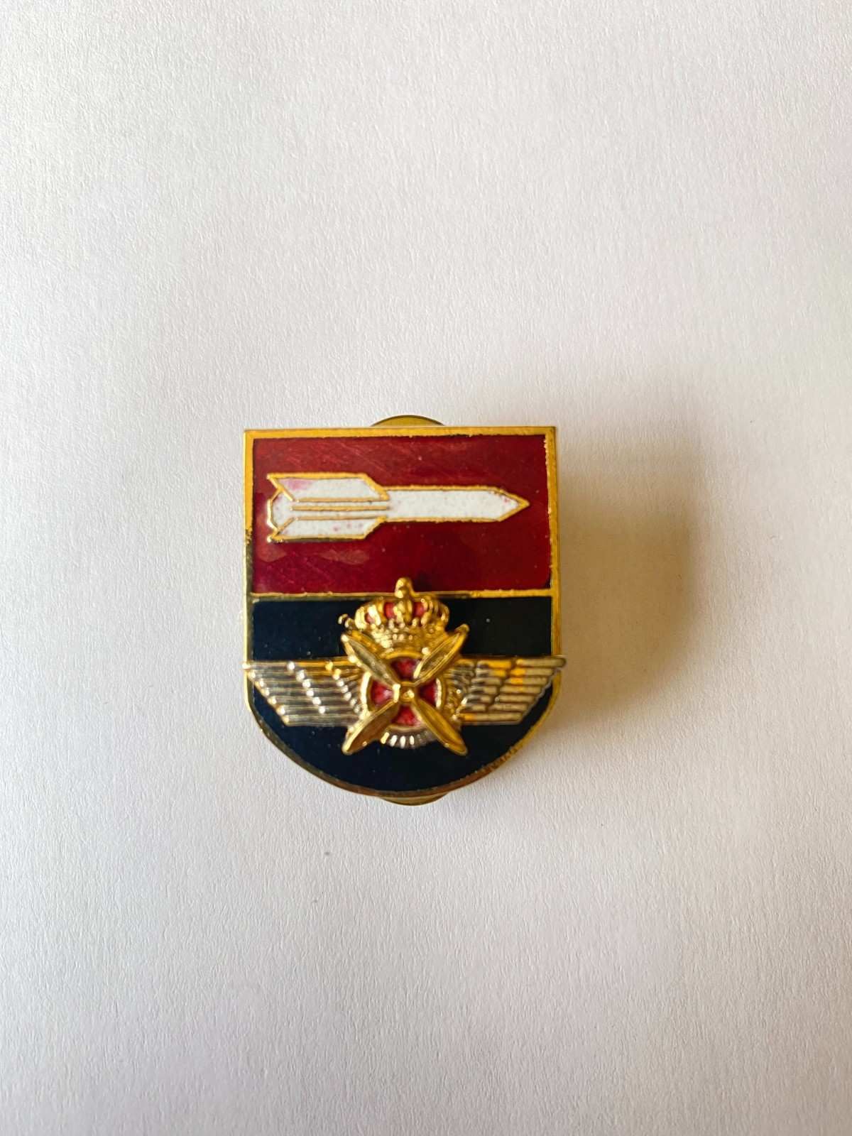 Pequeño broche con forma de escudo rojo y dorado con un emblema de espada y laurel sobre fondo blanco.