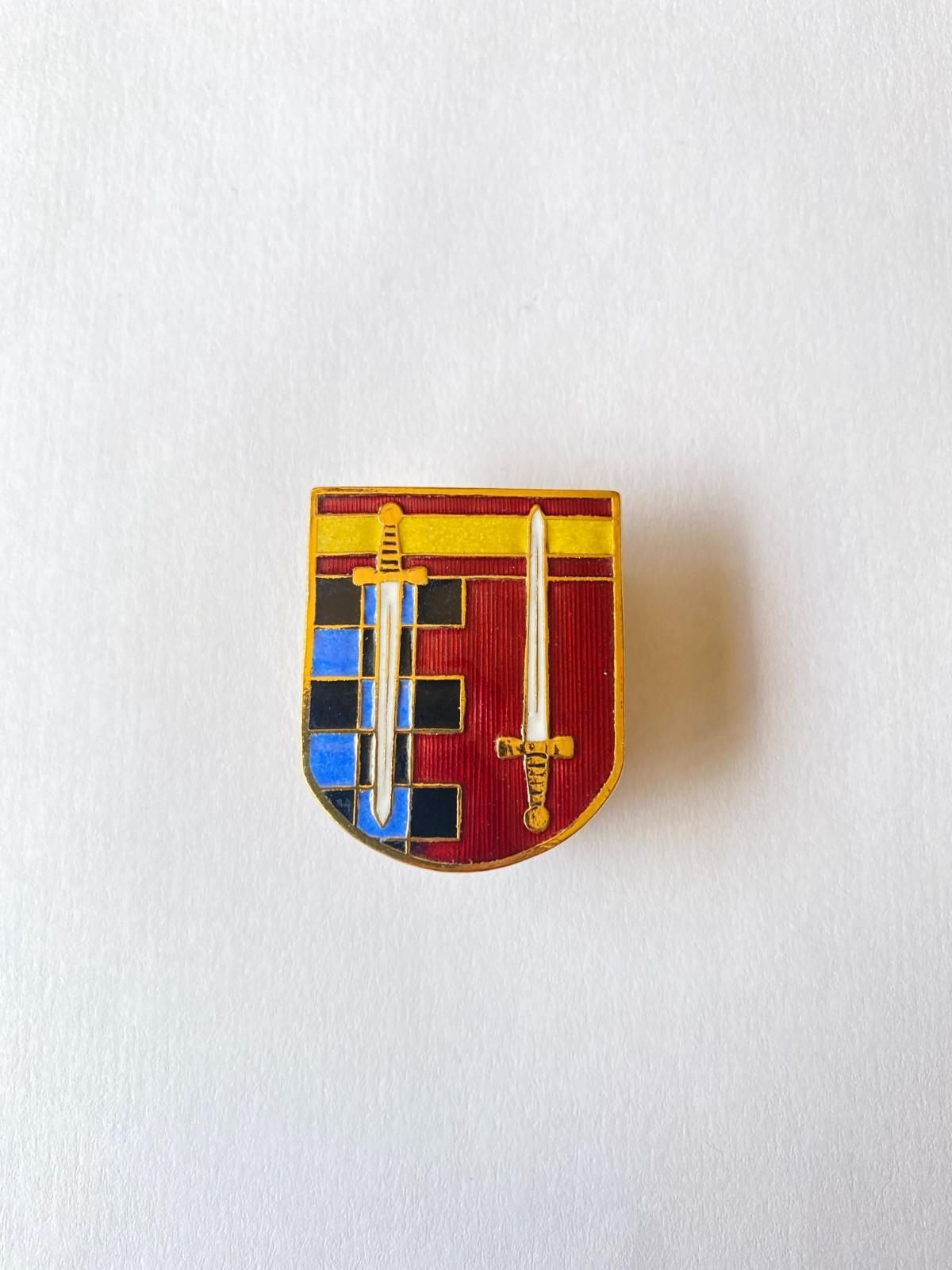 Pequeño broche con forma de escudo rojo y dorado con una cruz negra y cuadrados azules sobre fondo blanco.