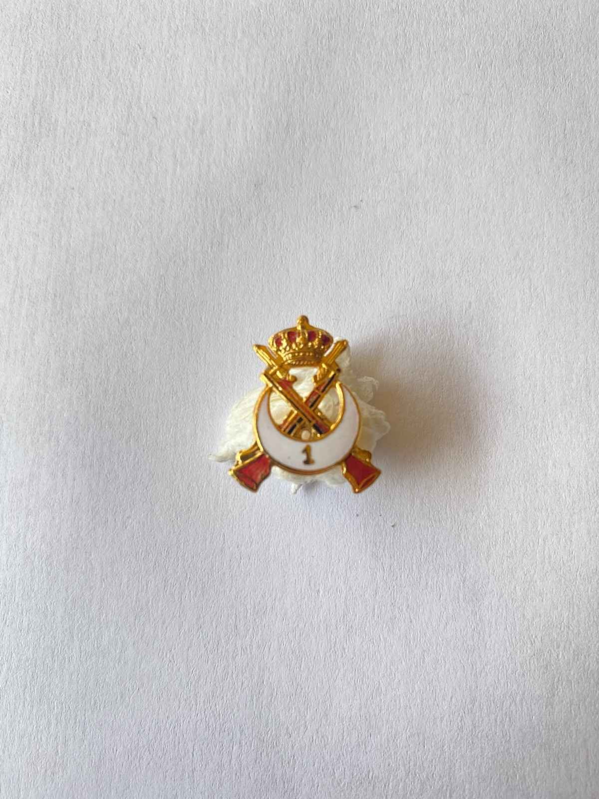 Pequeño broche de solapa dorado y blanco con detalles rojos sobre fondo gris claro.