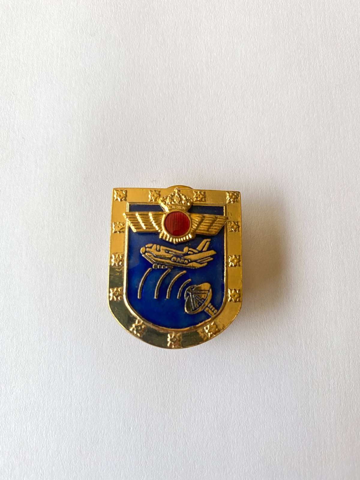 Pin con escudo azul y dorado con un emblema rojo sobre fondo blanco.
