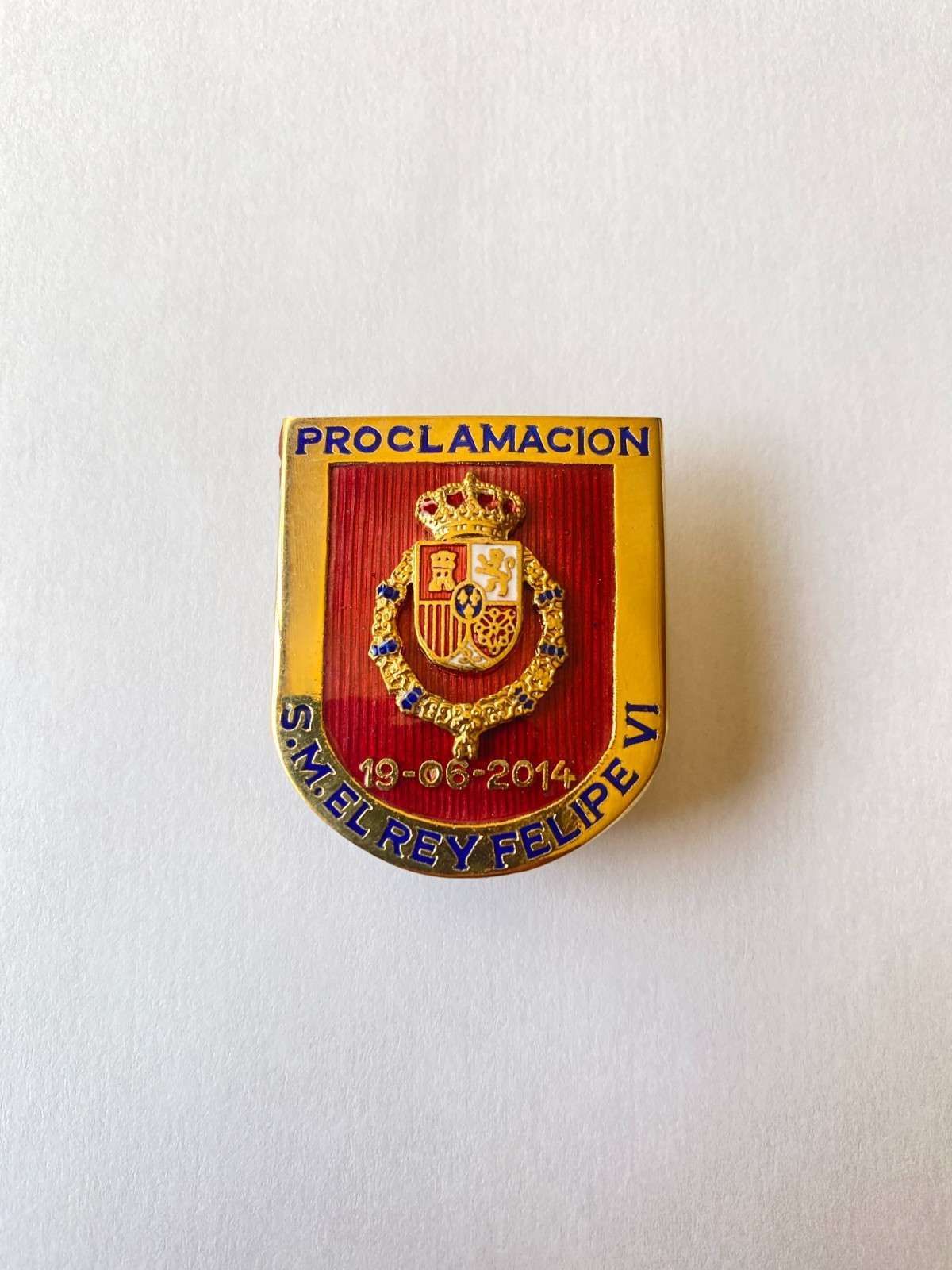 Insignia roja y dorada con forma de escudo, con la inscripción “PROCLAMACION” en la parte superior y un blasón en el centro.