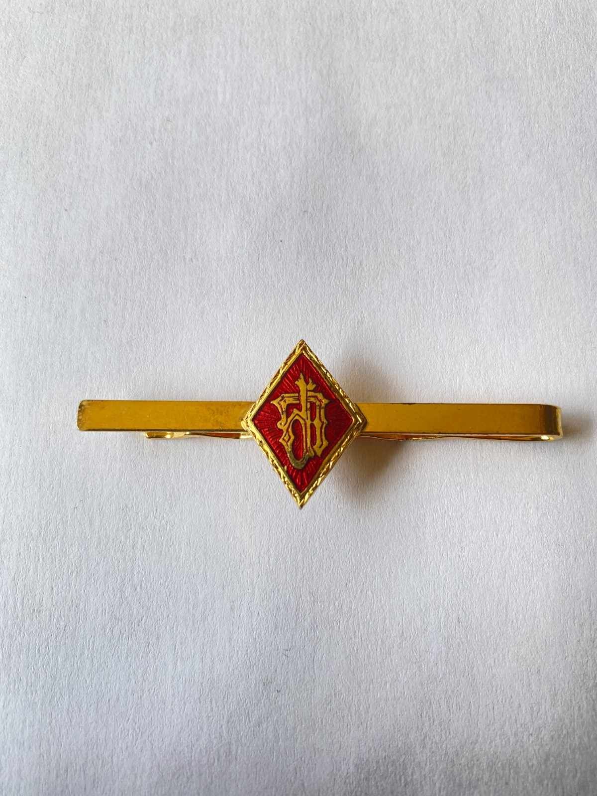 Pinza de corbata dorada con un emblema rojo en forma de diamante sobre fondo blanco.