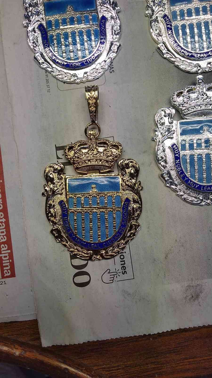 Medalla o colgante en forma de escudo azul y dorado expuesto entre varios trofeos de plata similares.