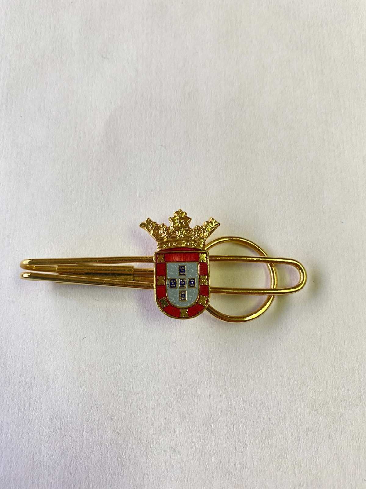 Pinza de corbata dorada con un emblema de escudo rojo y blanco sobre fondo claro.