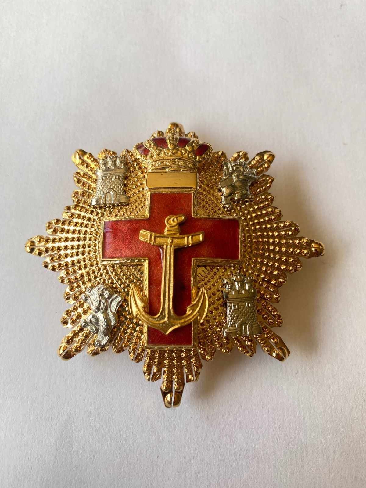 Insignia de cruz dorada con centro de esmalte rojo y una corona, sobre fondo blanco.