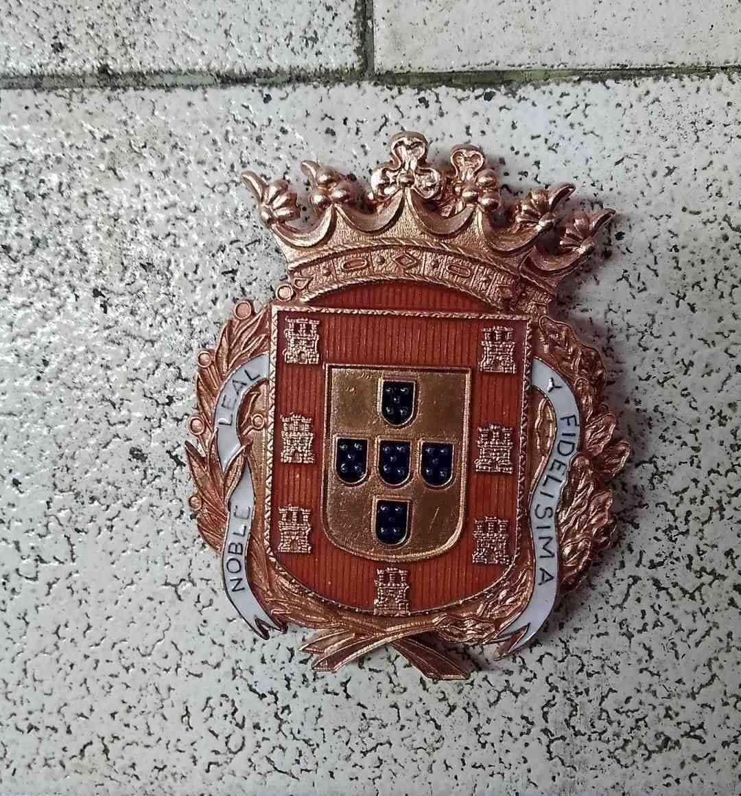 Escudo de armas portugués con corona sobre un muro de piedra gris.