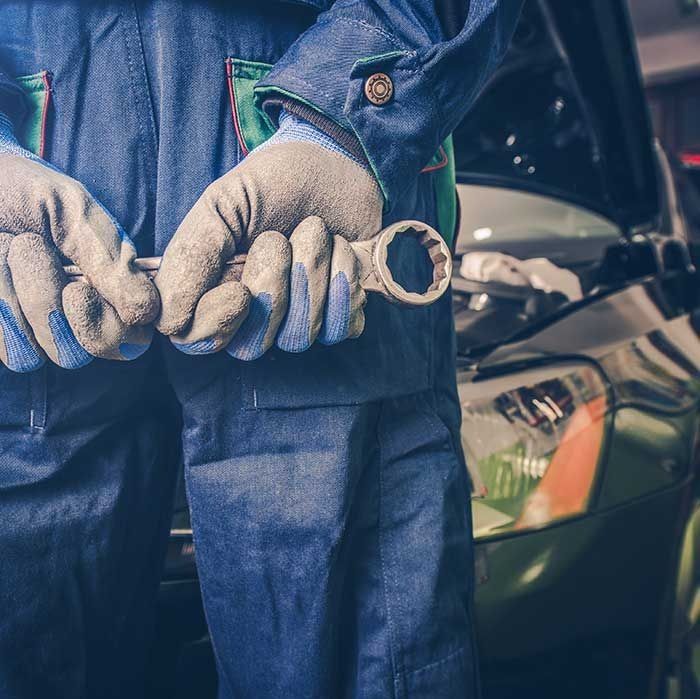 Mecánico con mono azul sosteniendo una llave inglesa, trabajando en un automóvil con el capó abierto.