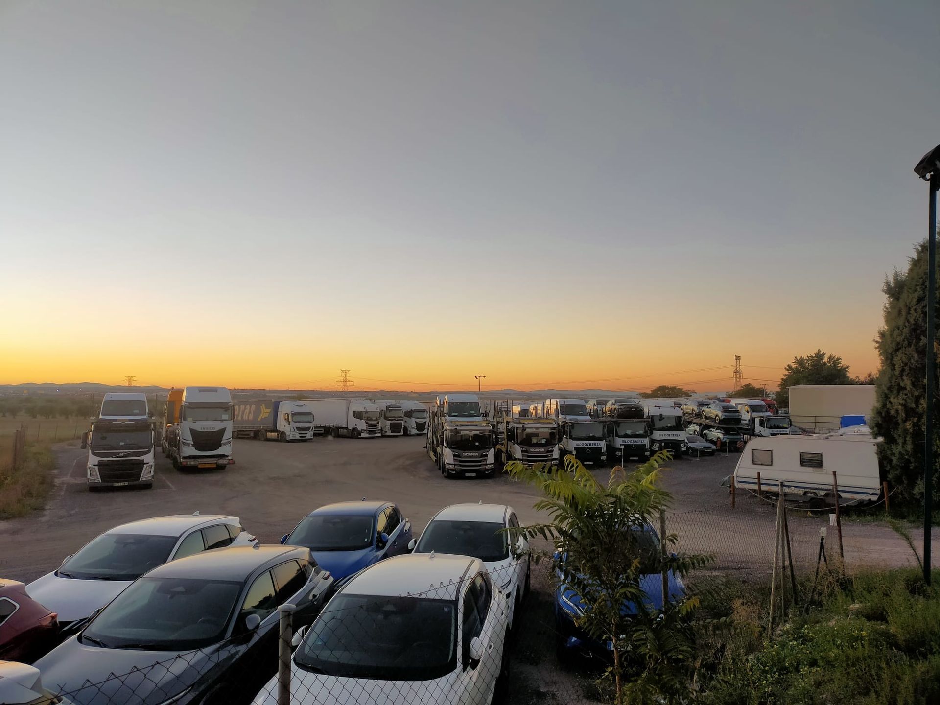 Un estacionamiento al atardecer con camiones semirremolque, automóviles y una caravana contra un cielo pálido.