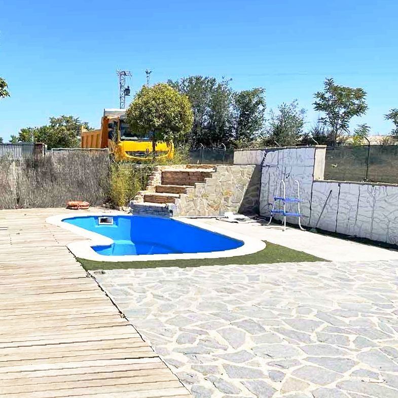 Piscina de agua azul rodeada de patio de piedra y deck de madera bajo un cielo azul claro.