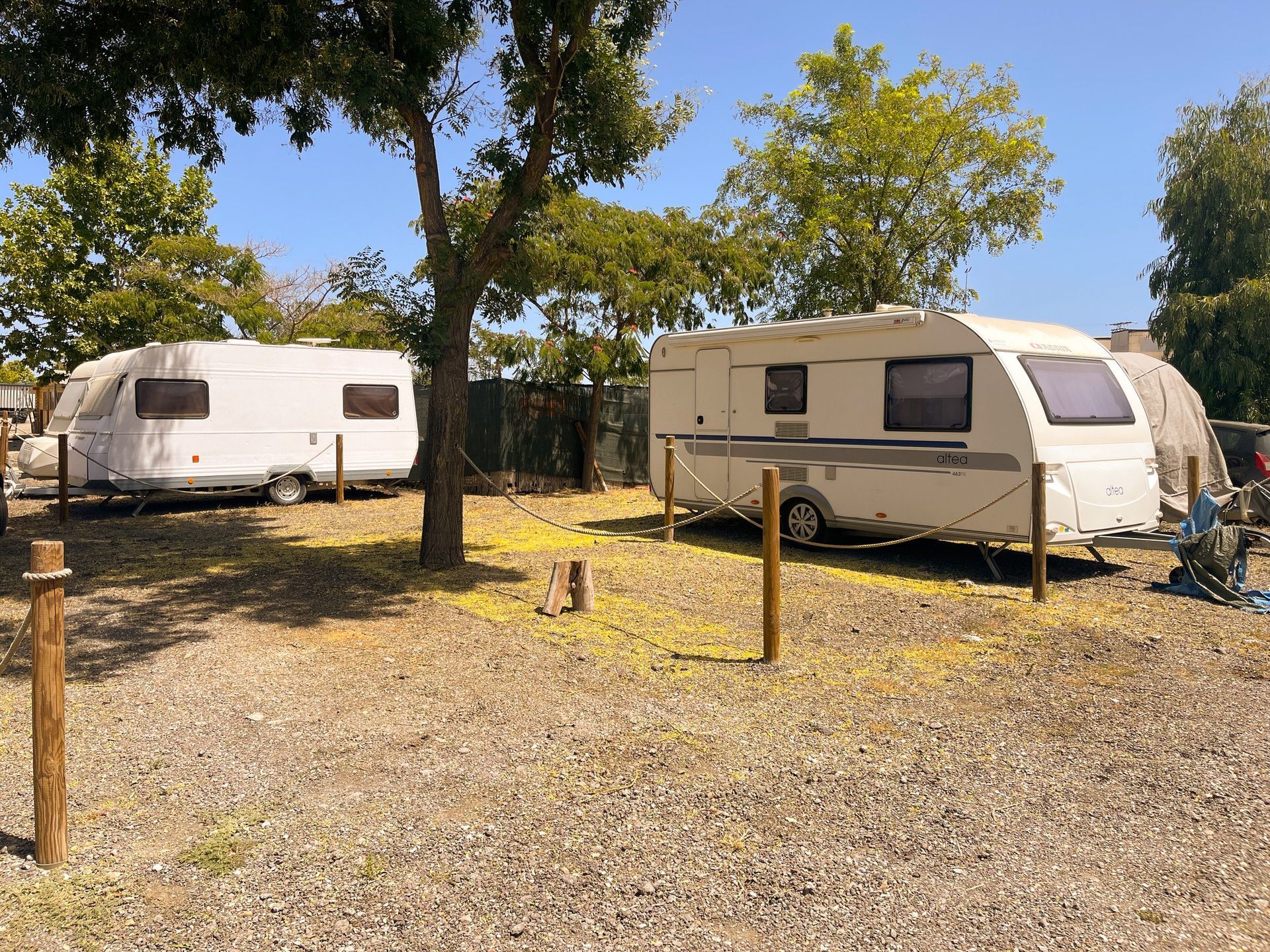Dos caravanas blancas estacionadas en un lote de grava bajo un cielo azul con árboles.