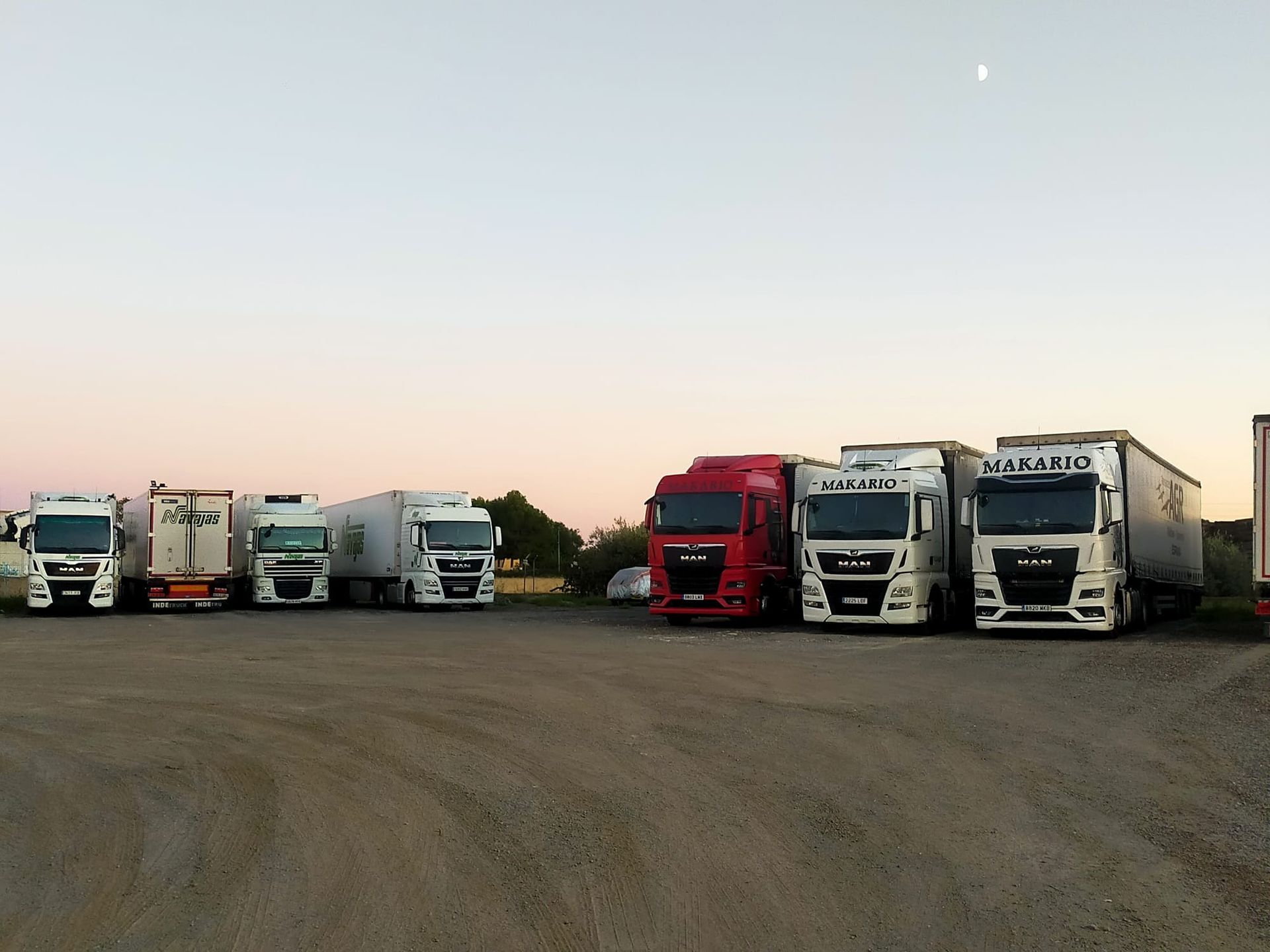 Camiones estacionados en un estacionamiento al anochecer. El cielo está rosado y se ve la luna.
