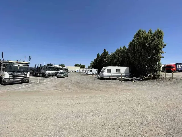 Terreno de grava con camiones, una caravana y árboles bajo un cielo azul brillante.