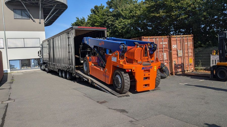 Ein orangefarbenes und blaues Industriefahrzeug wird auf einer Rampe in der Nähe von Schiffscontainern von einem Sattelzug entladen.