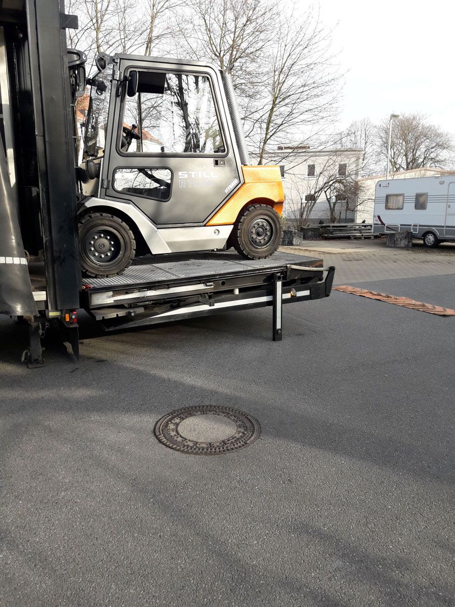Ein Gabelstapler steht auf einer Rampe, geparkt auf Asphalt neben einem Kanaldeckel. Im Hintergrund ist ein Fahrzeug zu sehen.