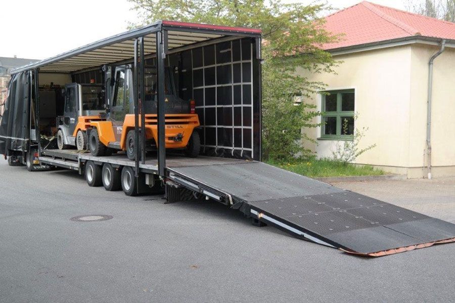 Gabelstapler auf einem Pritschenwagen mit abgesenkter Rampe zum Be- und Entladen vor einem Gebäude.