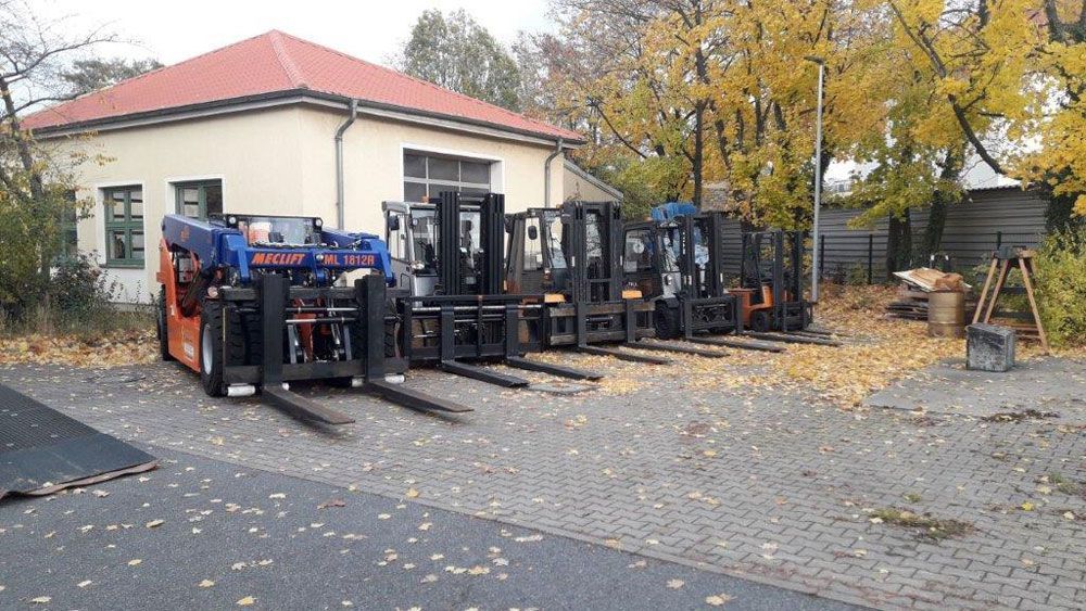 Gabelstapler stehen in einer Reihe vor einem Gebäude. Herbstlaub liegt auf dem Boden.