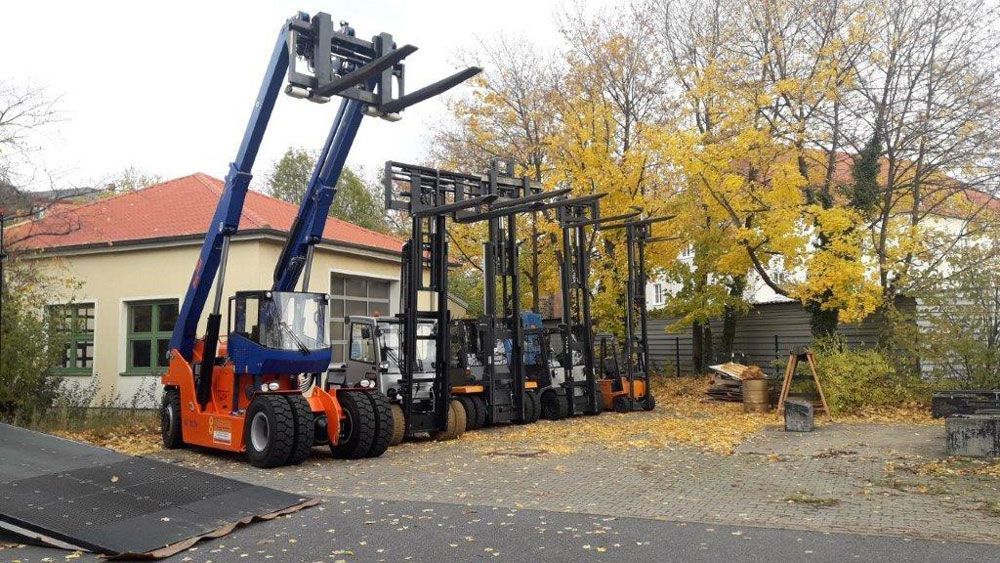 Oranger und blauer Gabelstapler mit ausgefahrenen Gabeln, geparkt neben anderen Gabelstaplern, Herbstkulisse.