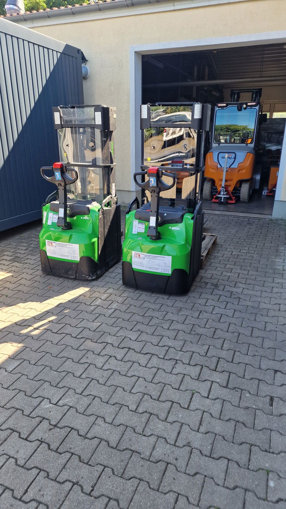 Zwei grüne elektrische Hubwagen stehen auf einer gepflasterten Fläche vor einer Garage; im Hintergrund ist eine weitere Maschine zu sehen.
