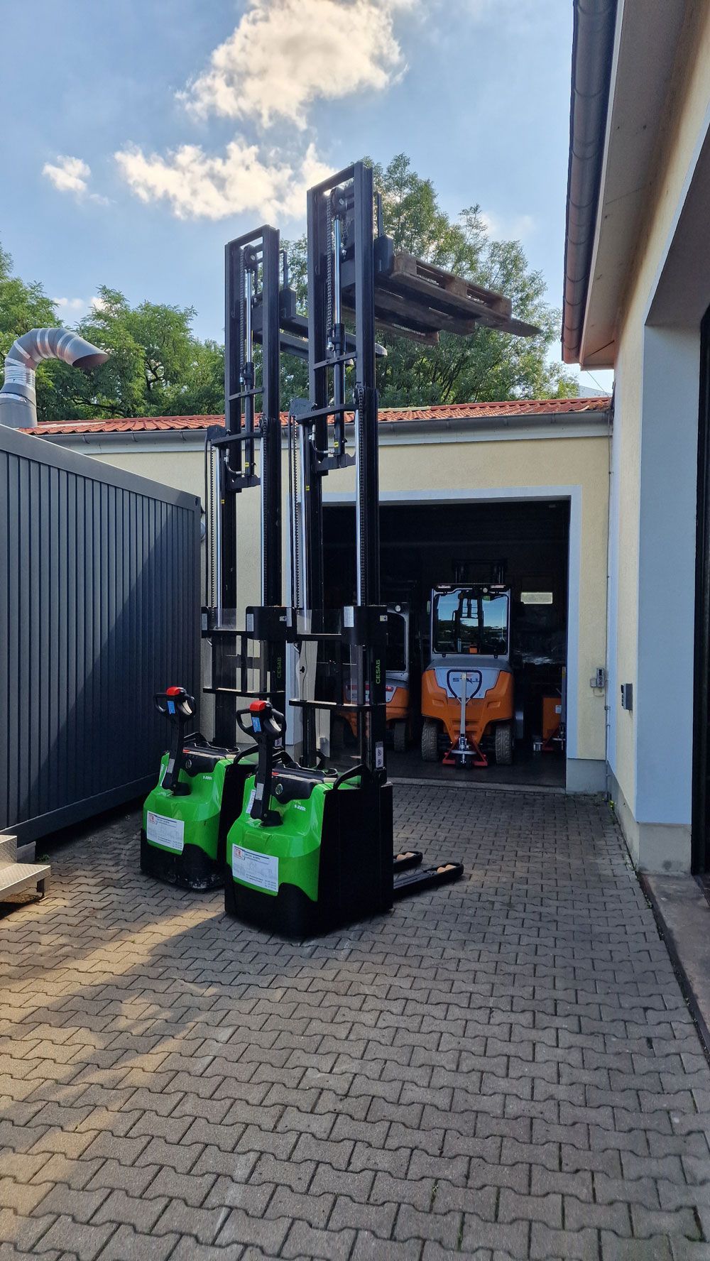 Zwei grüne und schwarze Elektrogabelstapler im Freien, in der Nähe eines Gebäudes und einer Garage, unter sonnigem Himmel.