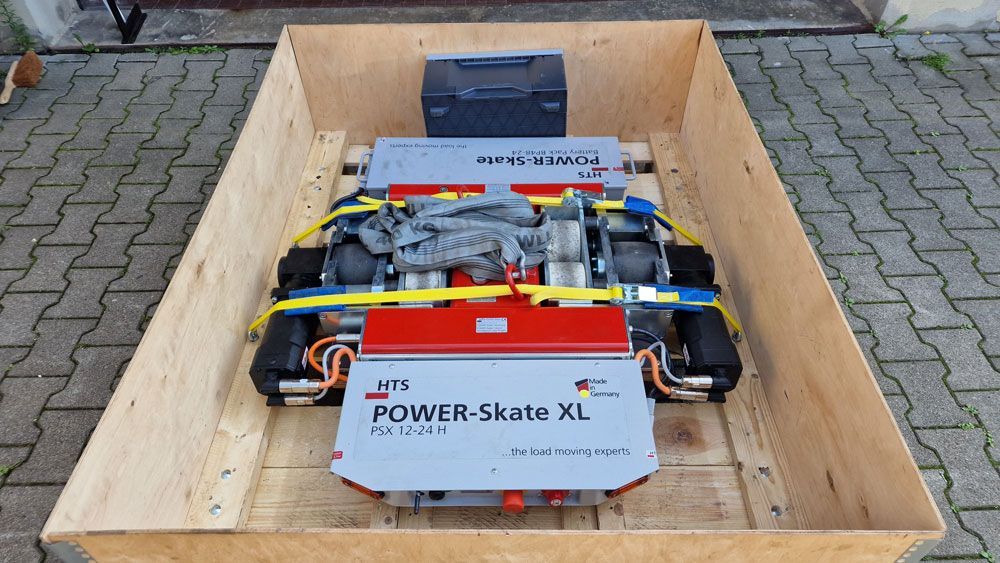Power Skate XL Roboter in einer Holzkiste, Vorderansicht. Rote, graue und schwarze Bauteile mit gelben Kabeln.