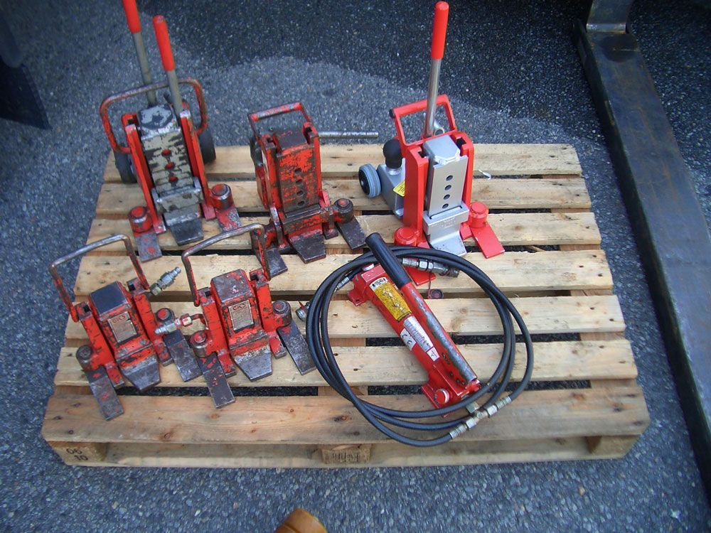 Mehrere rote Hydraulikheber auf einer Holzpalette; an einem Heber ist ein Schlauch angeschlossen.