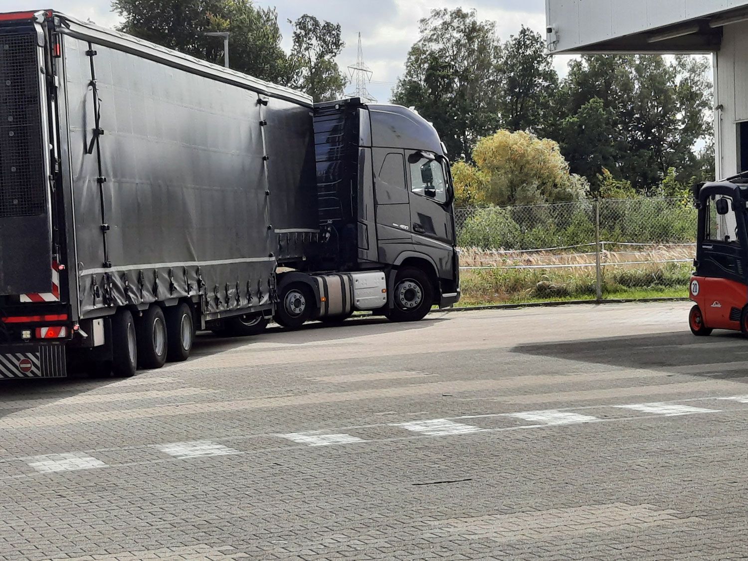 Ein Sattelzug steht an der Laderampe. Schwarzer Lkw, dunkler Anhänger, gepflasterter Boden, Gabelstapler in der Nähe.