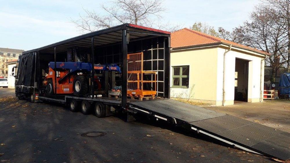 Ein großer LKW mit geöffneter Rampe, beladen mit Maschinen; neben einem kleinen Gebäude.