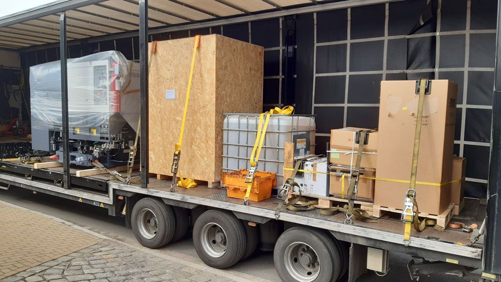 Die Ladefläche des Lastwagens ist mit großen Geräten und Kisten beladen, die mit gelben Gurten gesichert sind und für den Transport bereitstehen.