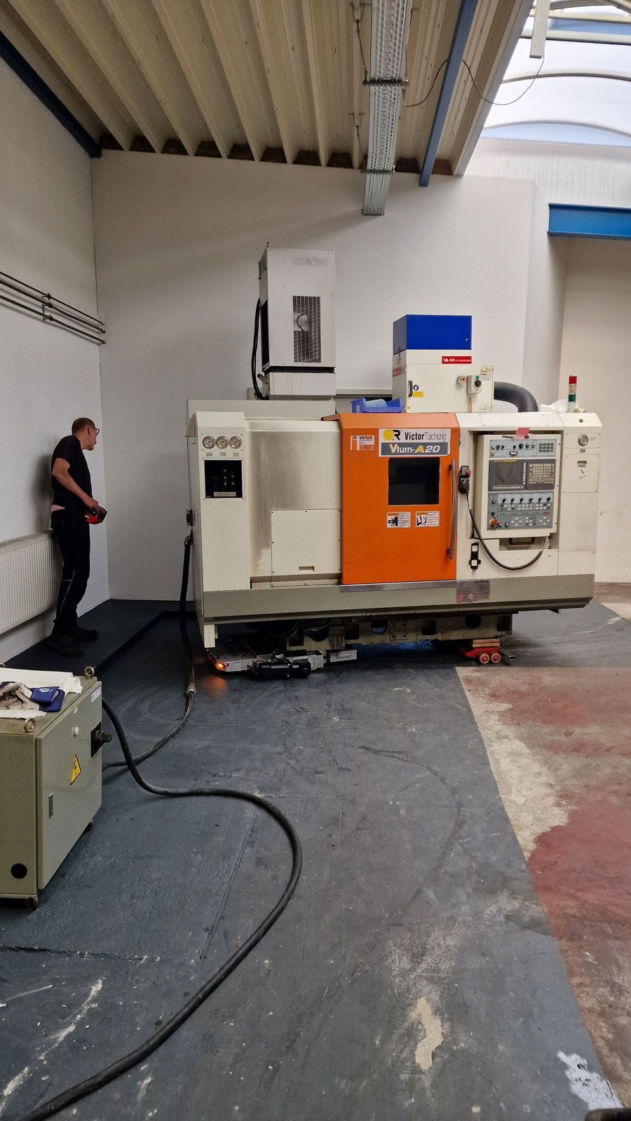 Ein Mann steht in einer Werkstatt neben einer industriellen CNC-Maschine. Die Maschine ist weiß, orange und blau.