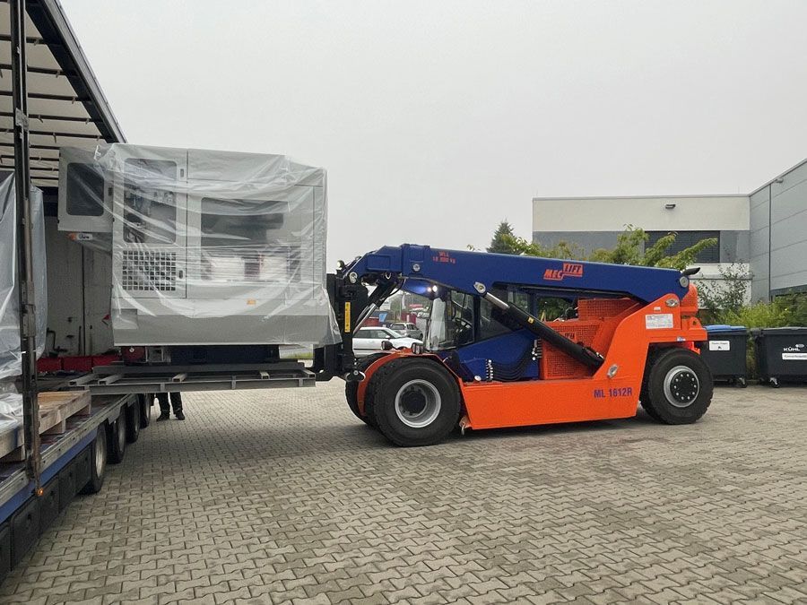 Ein orangefarbener und ein blauer Gabelstapler entladen Maschinen von einem LKW im Freien.