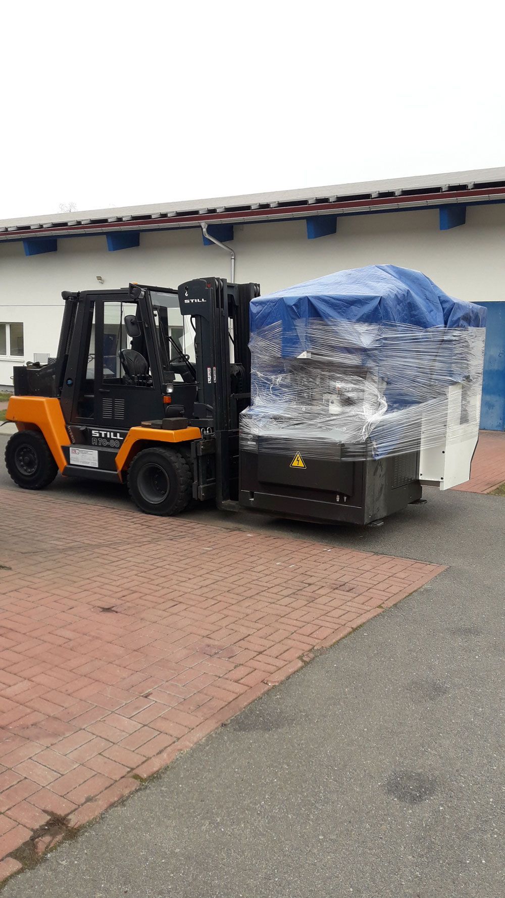 Ein Gabelstapler transportiert eine verpackte Maschine auf einem Ziegel- und Asphaltboden in der Nähe eines Gebäudes.