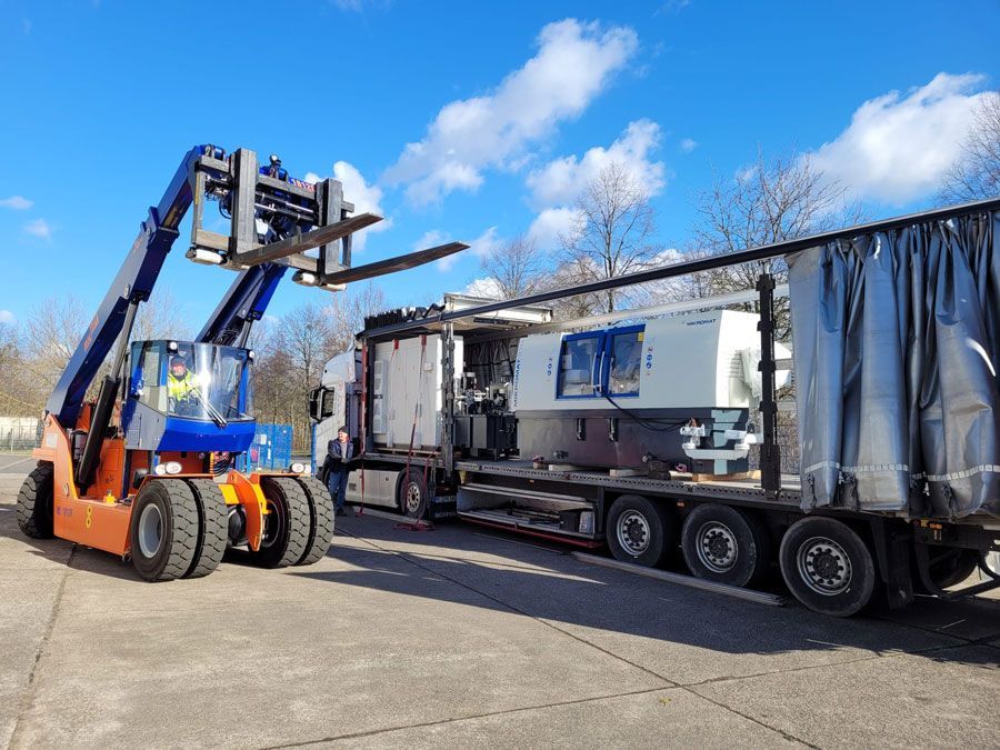 Gabelstapler verlädt Maschinen auf einen LKW. Oranger und blauer Gabelstapler, grauer LKW, Außenaufnahme, klarer Himmel.