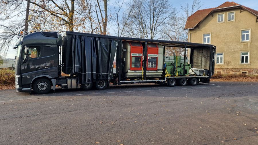 Ein schwarzer Sattelzug mit Industriemaschinen auf einem Pritschenauflieger parkt auf Asphalt in der Nähe eines Gebäudes.