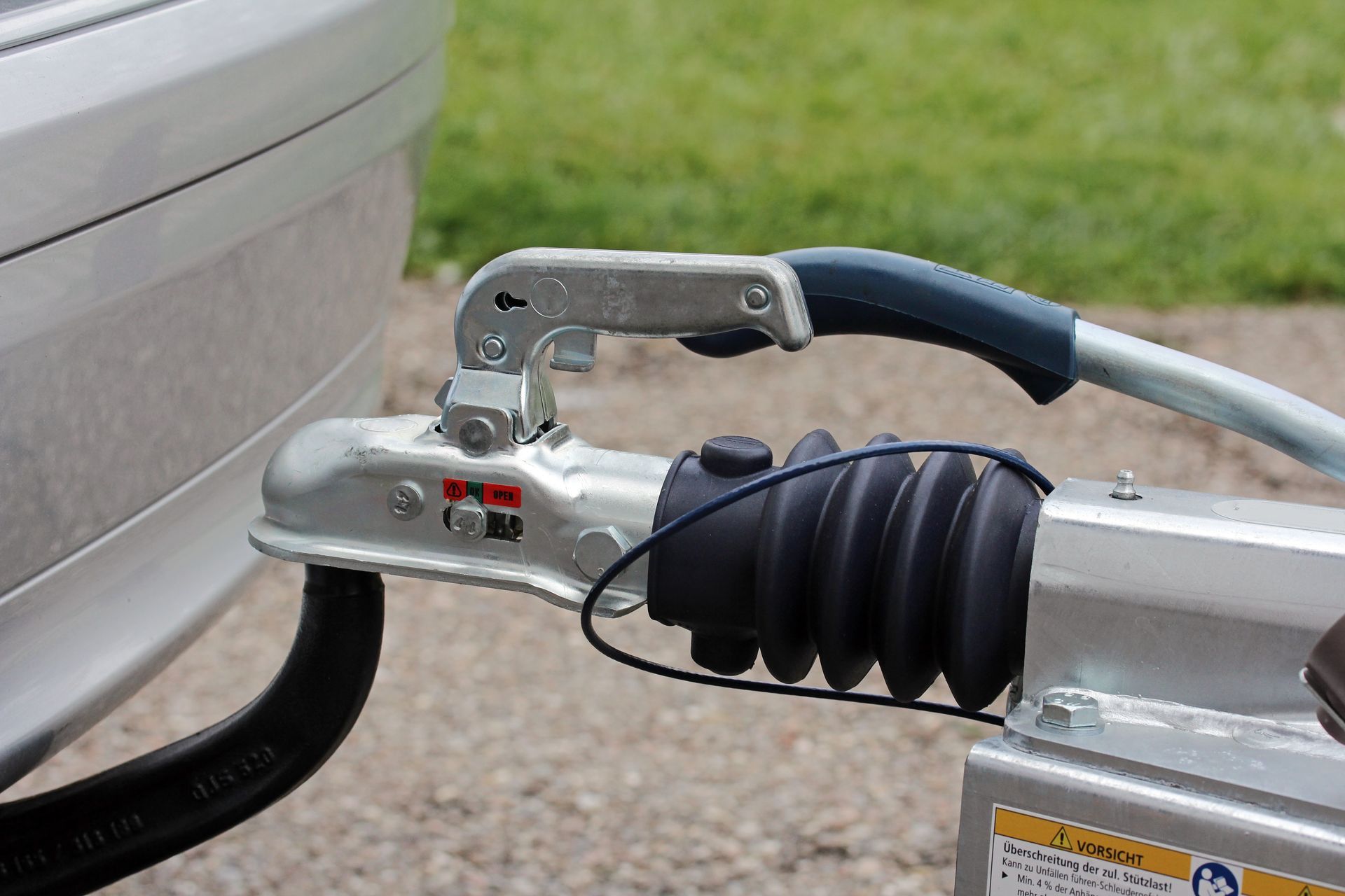 Un attelage de remorque fixé à une voiture. Il est argenté avec un soufflet en caoutchouc noir. La poignée est relevée.