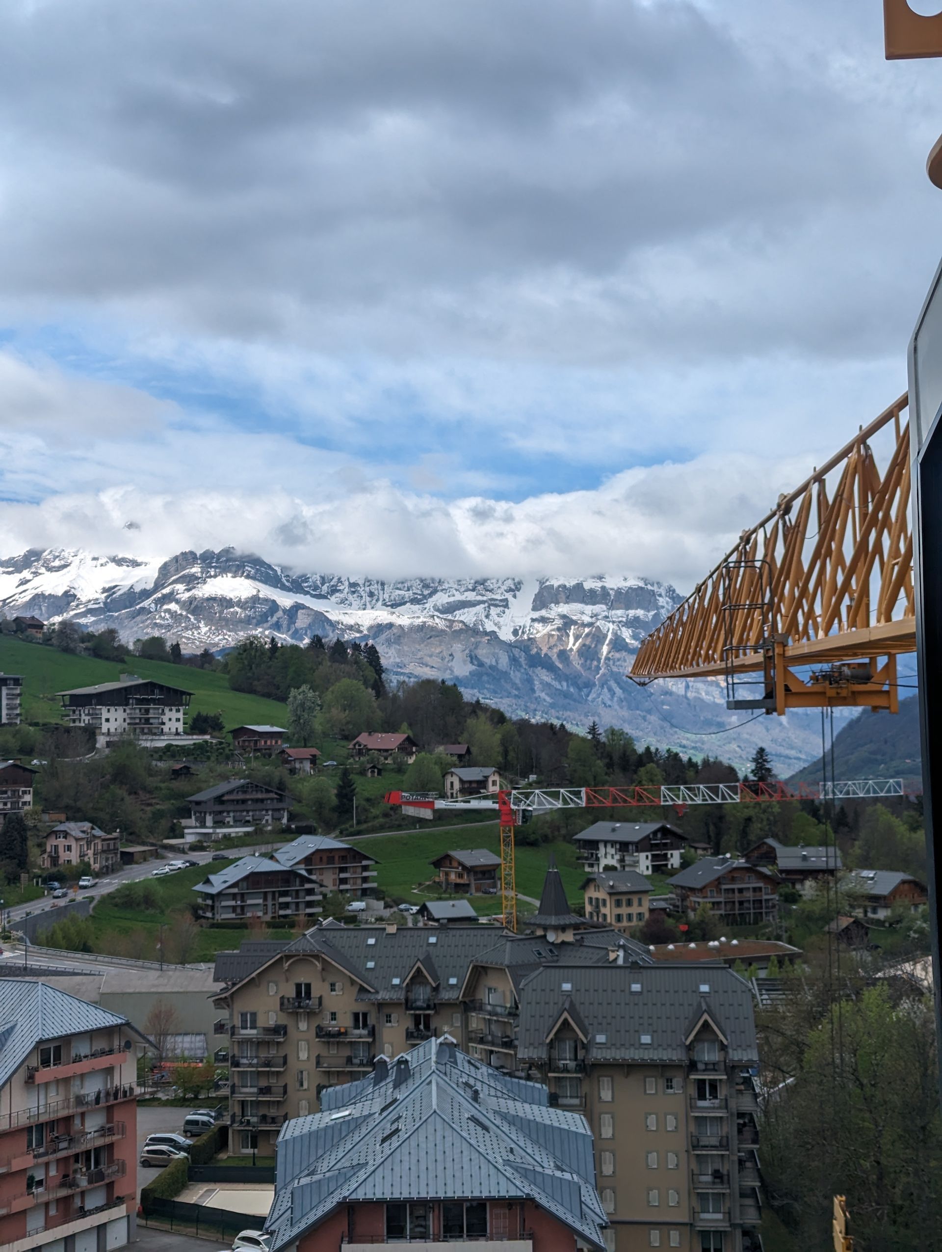 Paysage urbain avec des montagnes enneigées en arrière-plan, ciel nuageux. Maisons et grue de chantier visibles.