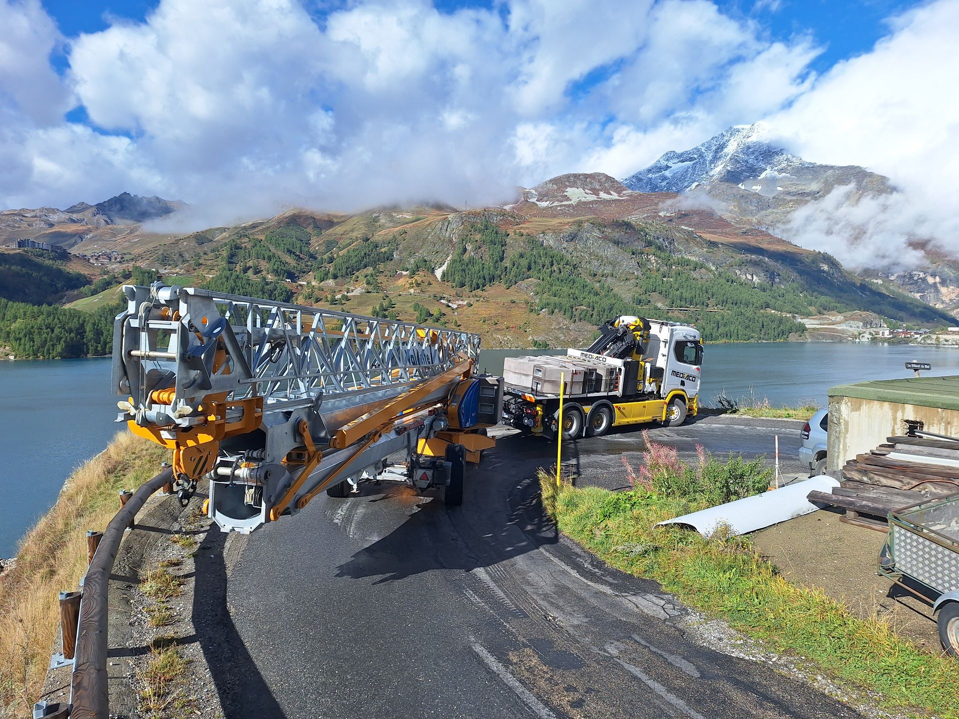 Des engins de chantier déchargent des matériaux près d'un lac et de montagnes.