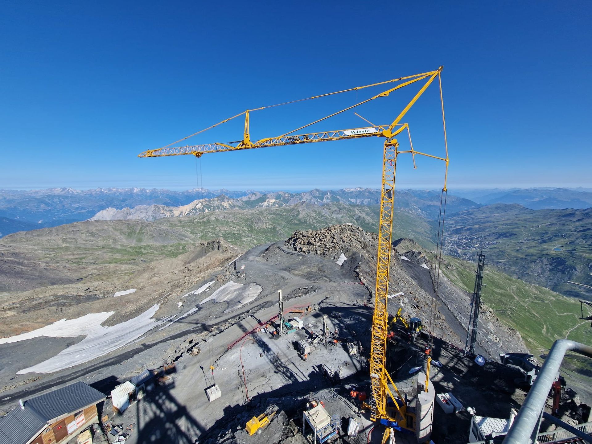 Grue jaune au sommet d'une montagne, chantier visible. Terrain rocailleux, montagnes en arrière-plan, ciel bleu.