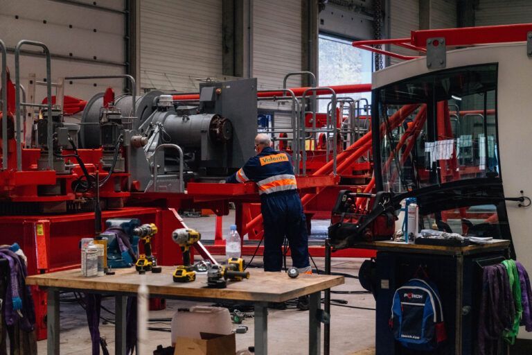 Un mécanicien en combinaison bleue travaille sur des machines dans un atelier équipé de matériel rouge et gris.