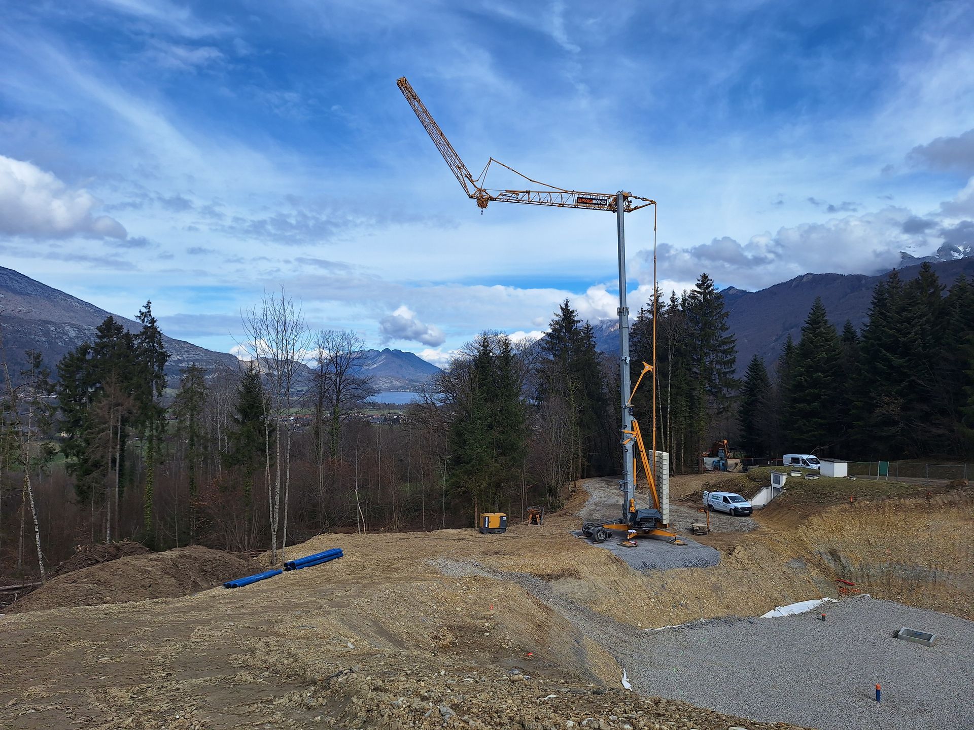 Chantier de construction en milieu montagneux avec une grue, des engins de chantier, un terrain nu, des arbres, un lac et un ciel nuageux.