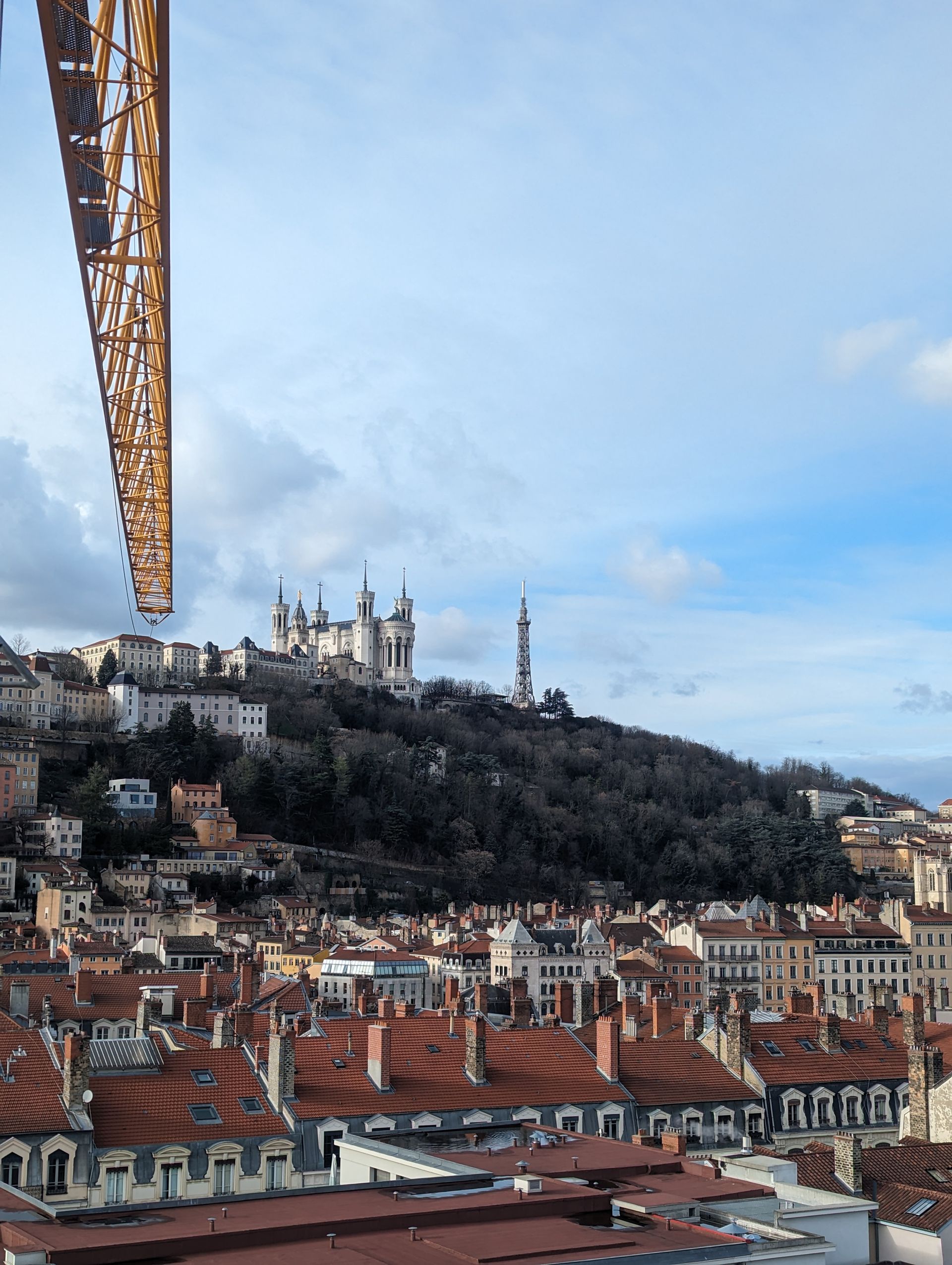 Vue de Lyon, en France, avec la basilique au sommet d'une colline et une grue de chantier au premier plan, sous un ciel bleu.