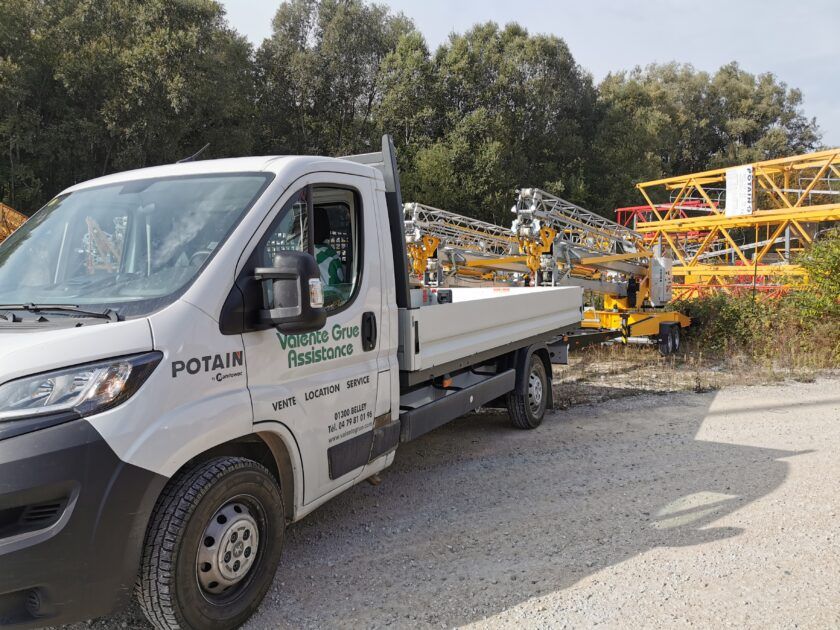 Camion blanc portant le logo Potain transportant des pièces de grue sur un chemin de gravier en bordure de forêt.