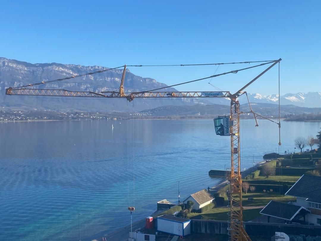 Grue de chantier au-dessus d'un lac, avec des montagnes en arrière-plan, sous un ciel bleu.