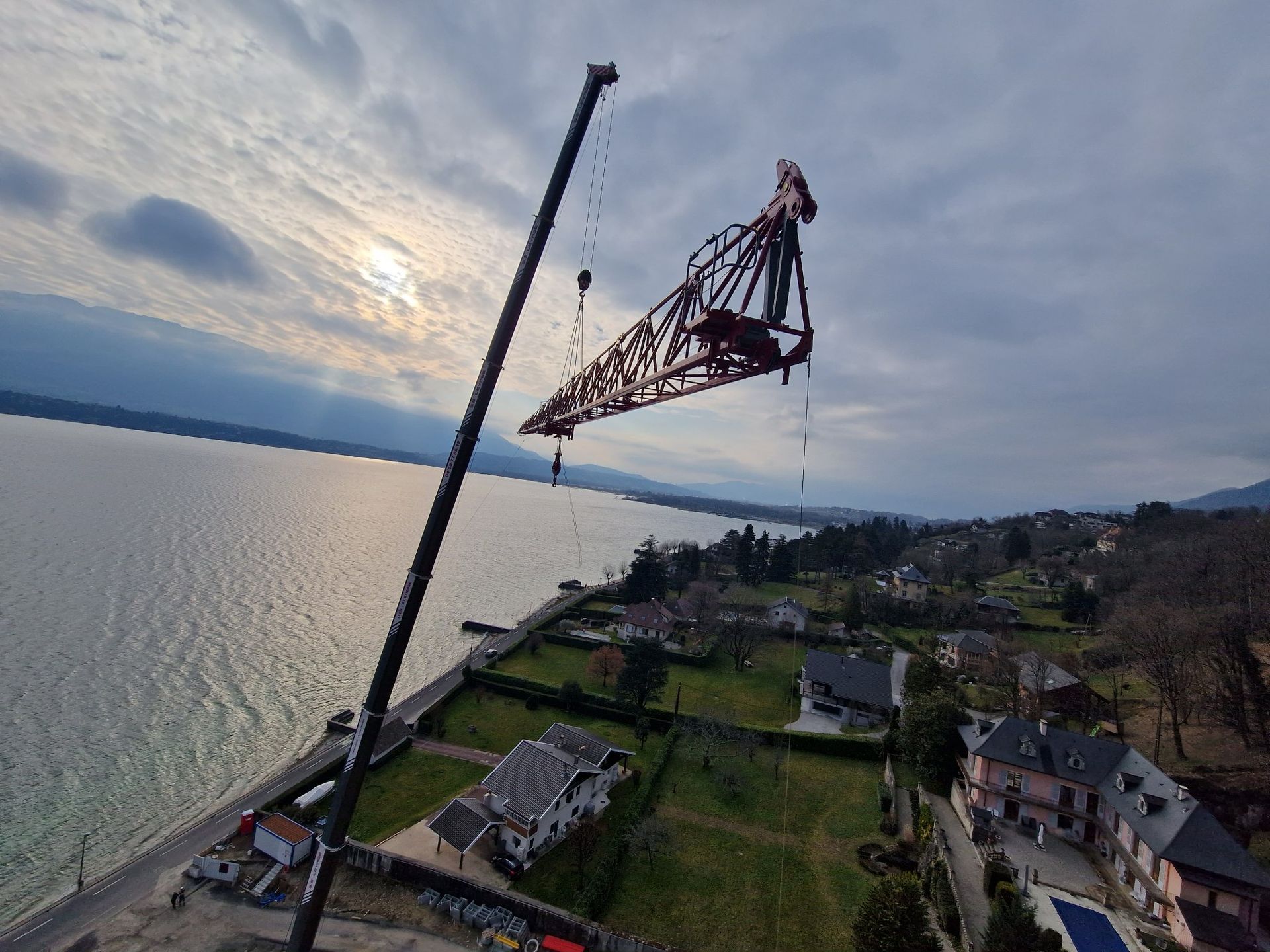 Une grue soulève une grande structure métallique au-dessus d'une étendue d'eau et d'un rivage bordé de maisons. Ciel nuageux.